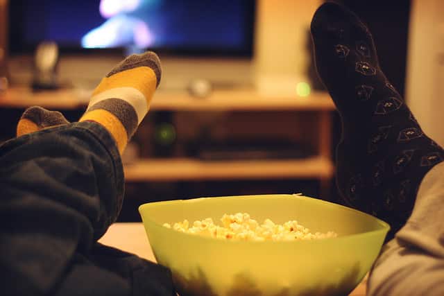 A bowl of popcorn in between 2 peoples's legs, who are lying back watching TV